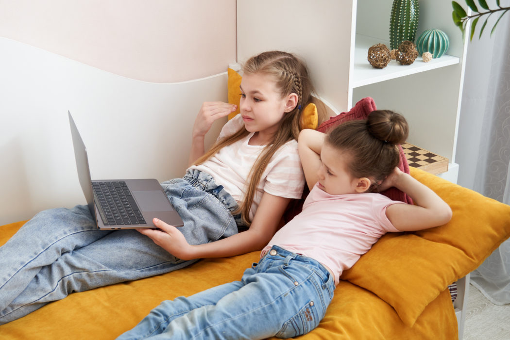 Two sisters lying on a bed together using laptop watching something